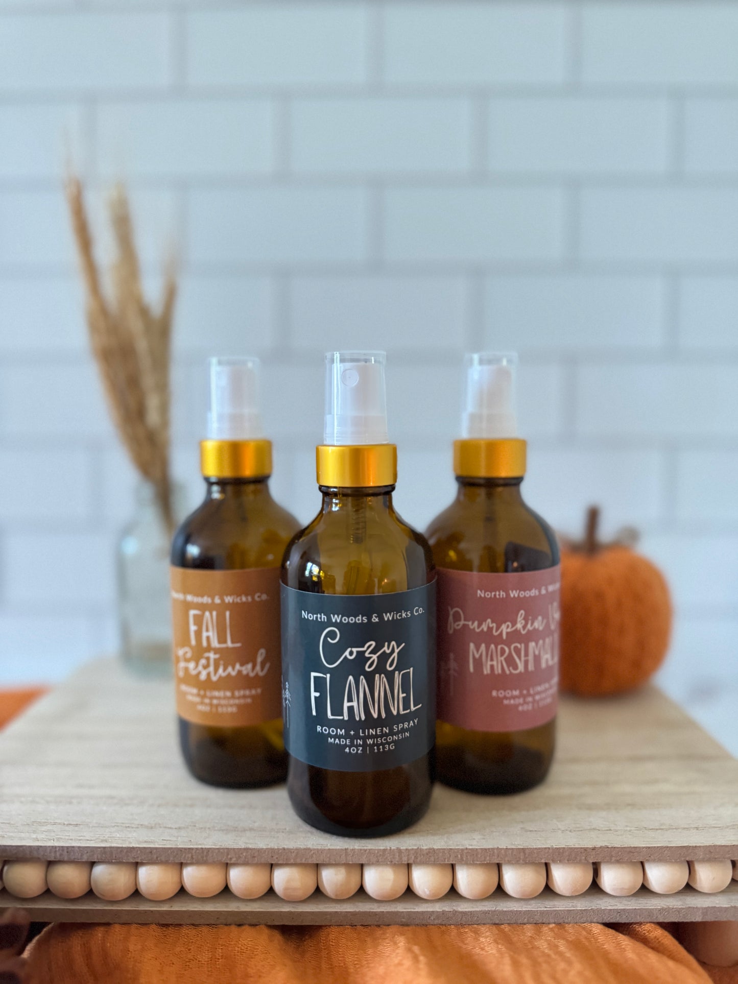 Three bottles of North Woods & Woods Co. room sprays on a wooden surface with a white tiled wall background.