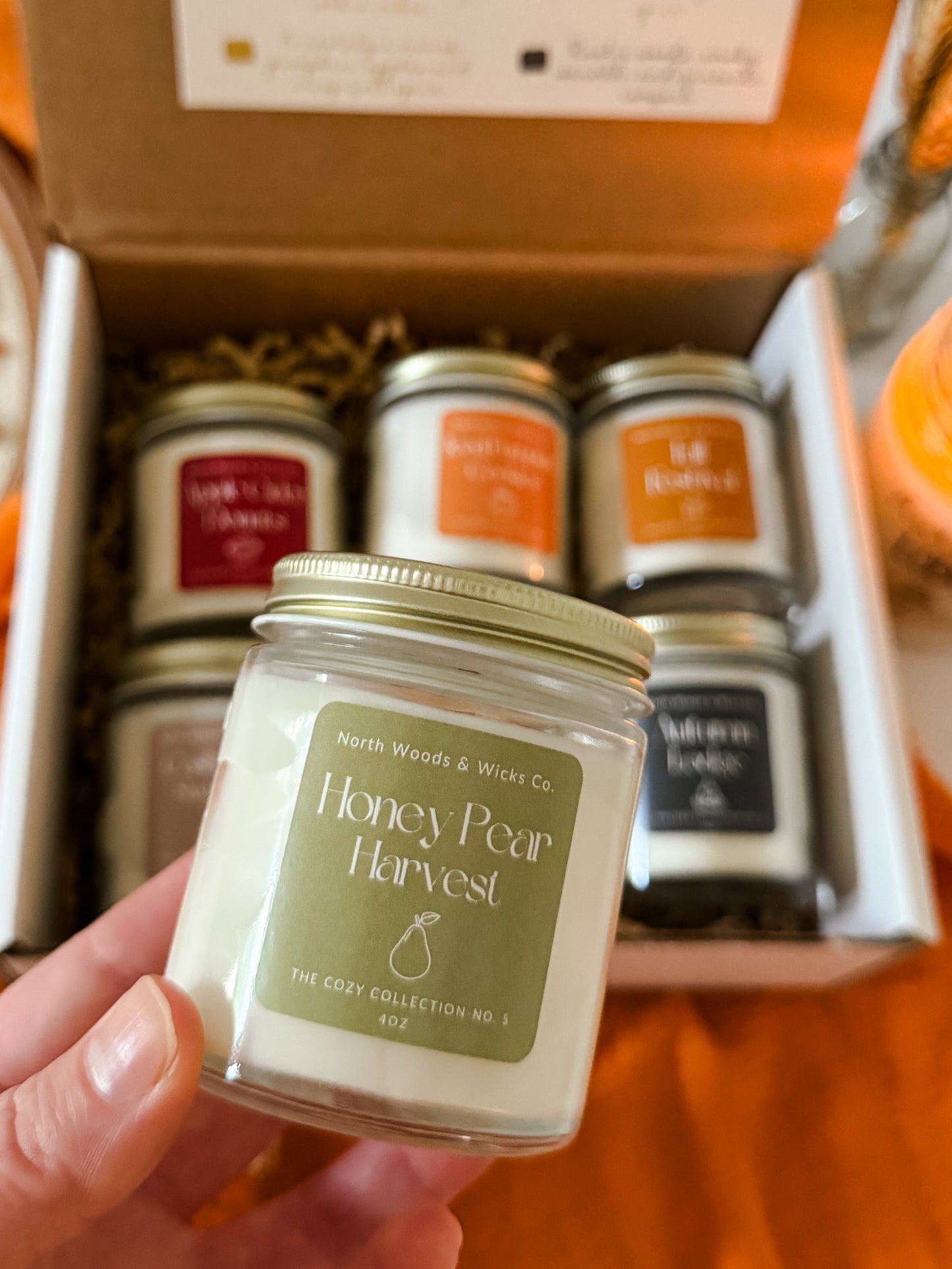 Candle labeled 'Honey Pear Harvest' held in front of a box containing other candles.