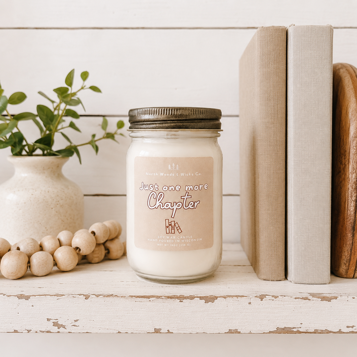 A glass jar candle with a metal lid and a beige label reading 'Just one more Chapter' sits on a rustic white wooden surface. Decorative books, a small vase with greenery, and wooden beads surround it in a styled home setting.